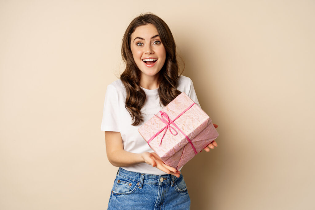 Home celebration and holidays concept. happy young woman holding gift wrapped in pink box, receive present, looking amazed and surprised, standing over beige background